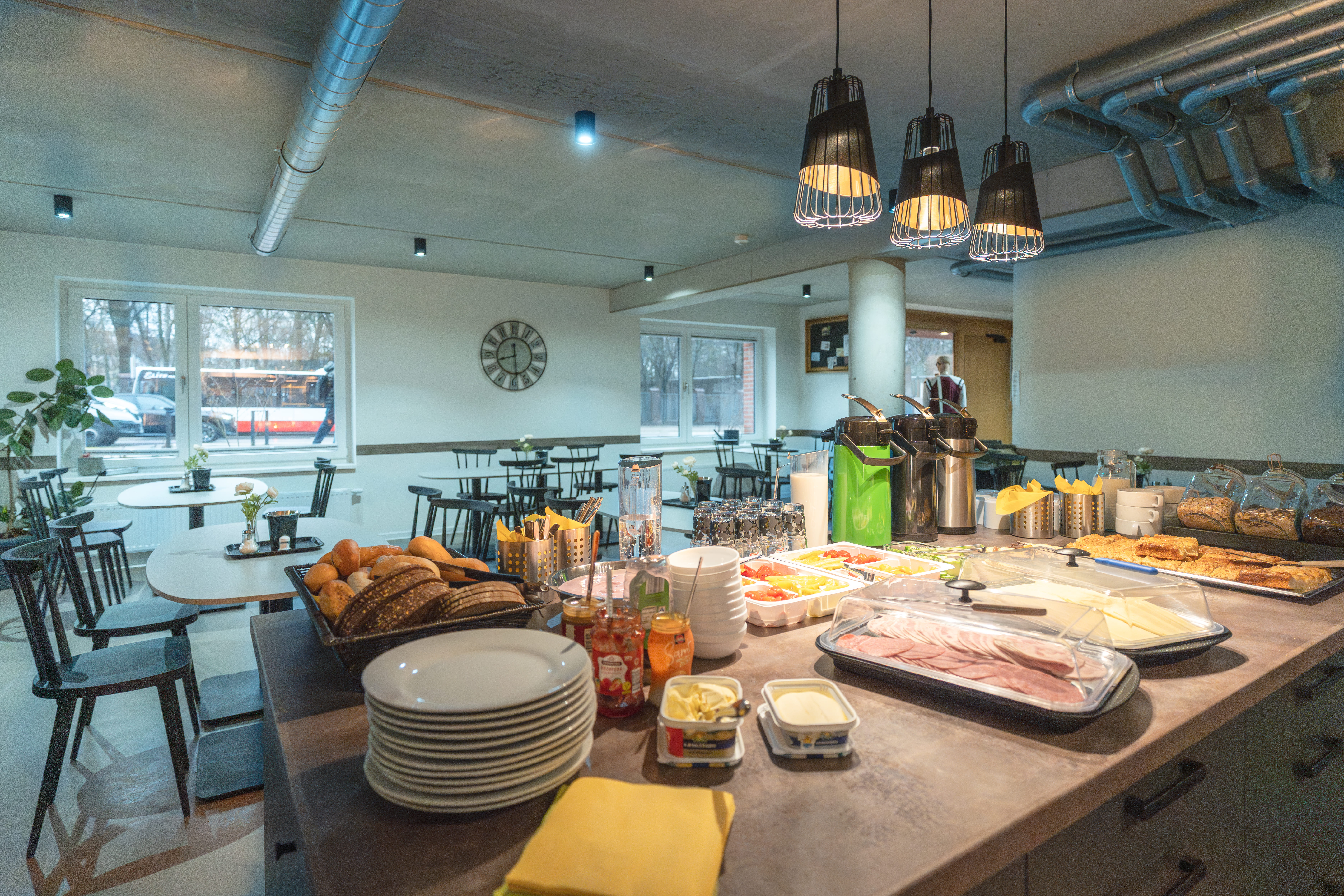 Cafeteria mit Buffet aus Brot, Aufschnitt, Käse und Getränken; industrielle Deckenlampen hängen über Tischen und Stühlen.