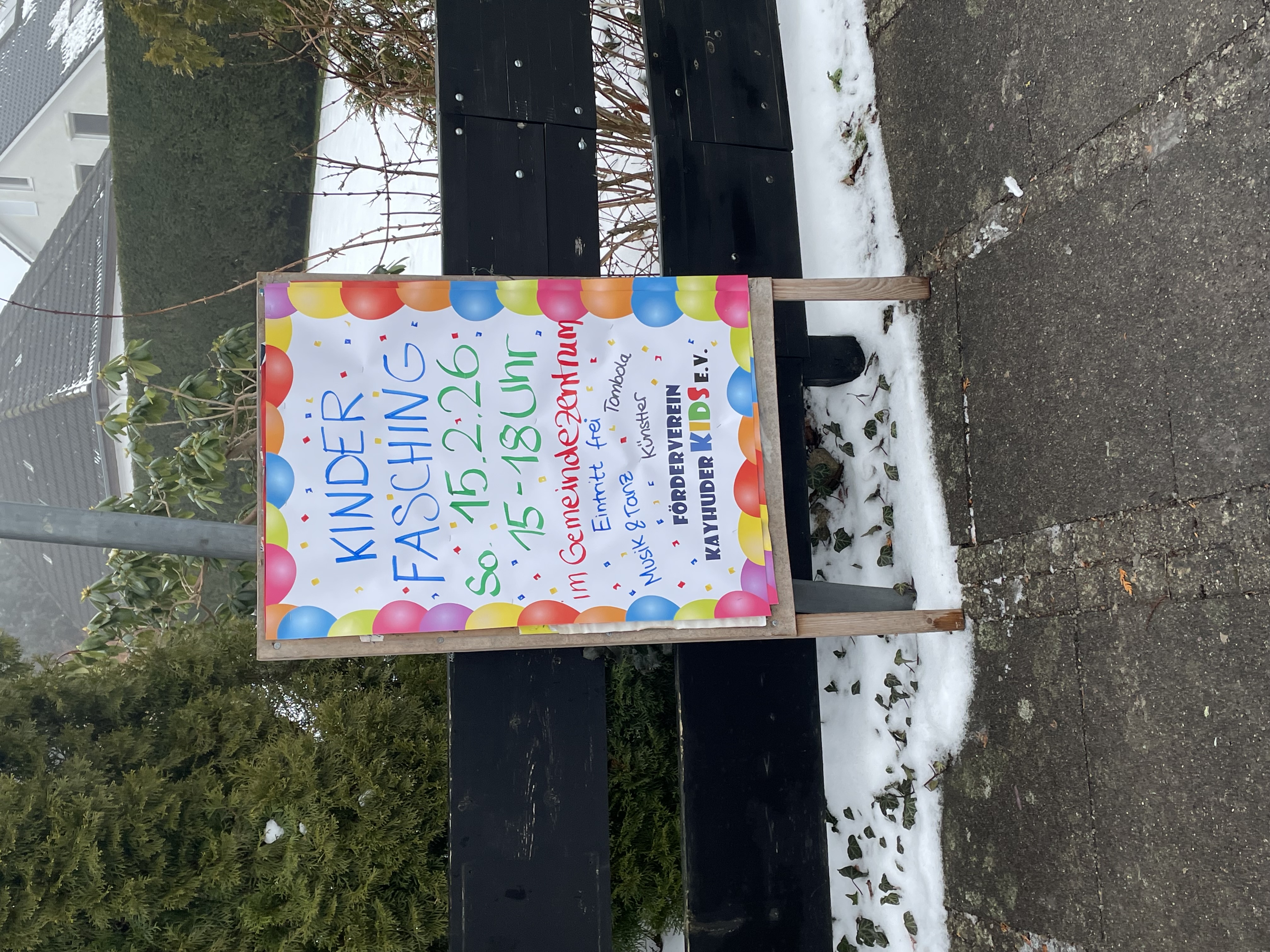 Farbiges Plakat „Kinderfasching“ mit Ballonrand, Datum 15.2.26, 15–18 Uhr, am Gemeindezentrum, vor schwarzem Zaun.