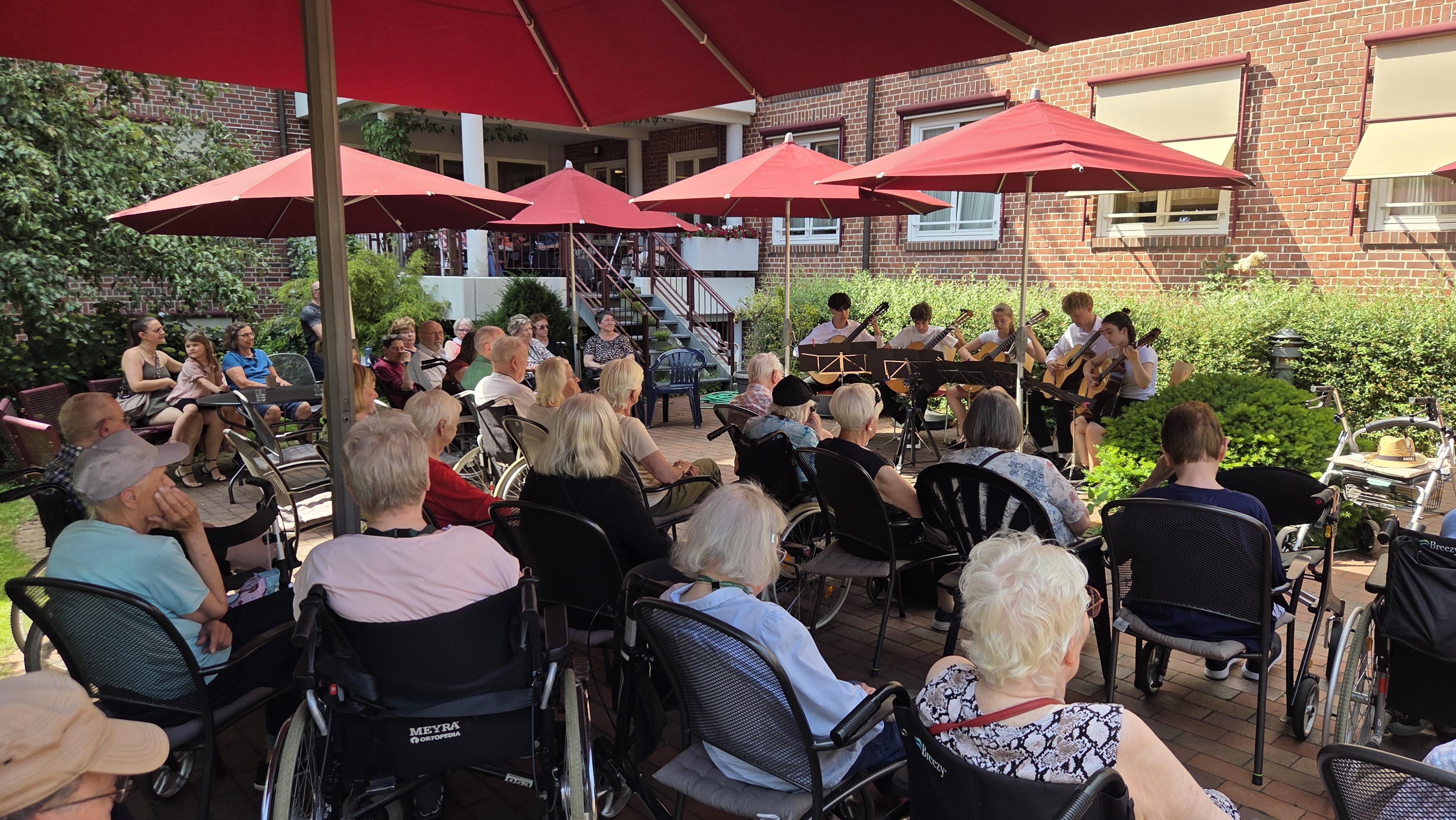 Gitarrengruppe spielt vor Publikum älterer Menschen auf einer Terrasse mit roten Sonnenschirmen vor einem Backsteingebäude.