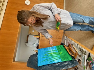 Eine Frau malt mit Pinsel und Palette eine grüne Graslandschaft auf einer Staffelei.