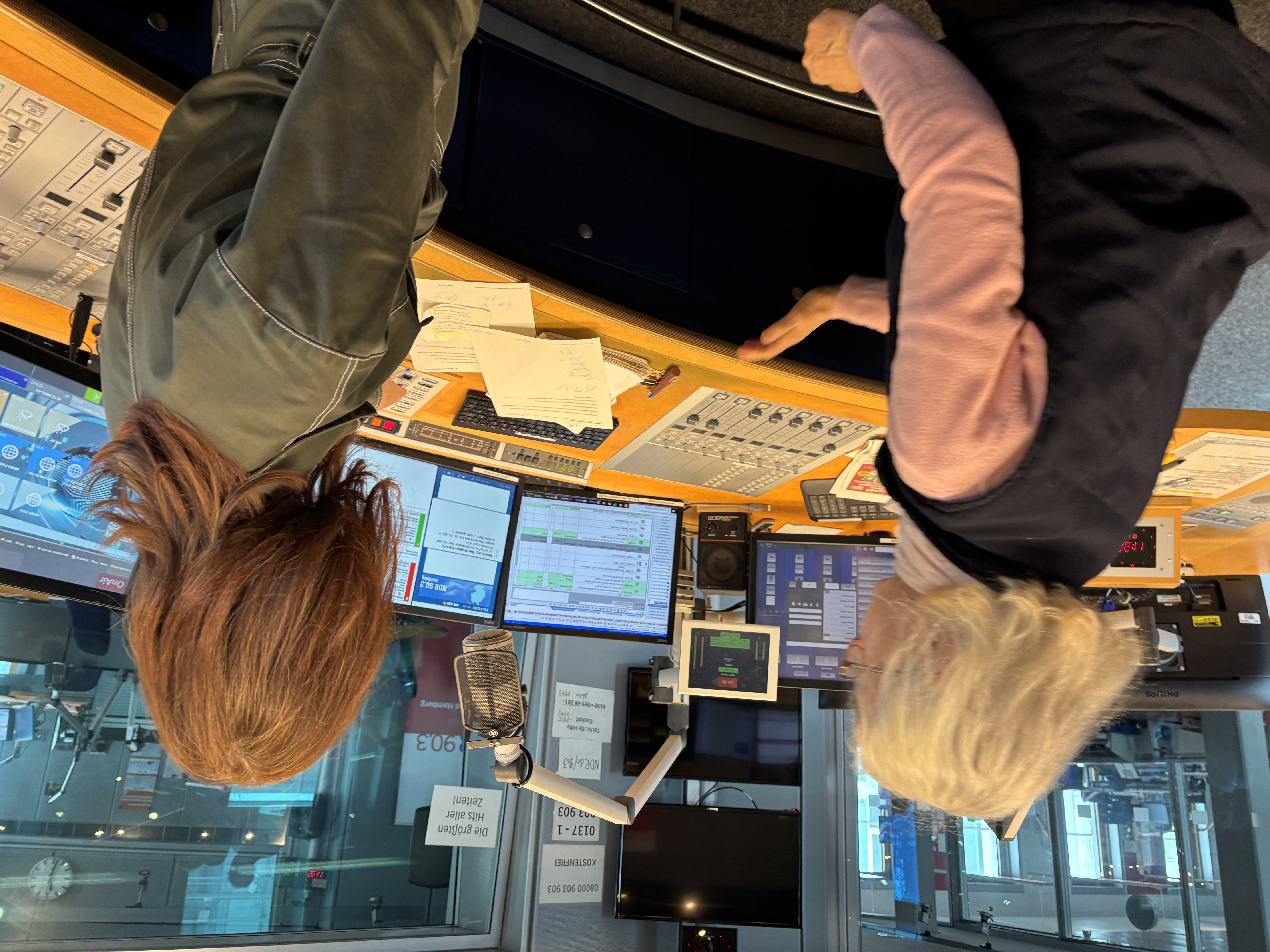 Zwei Radiomoderatoren sitzen an Mischpult und Monitoren in einem Sendestudio, Blick durch Glas auf andere Räume.