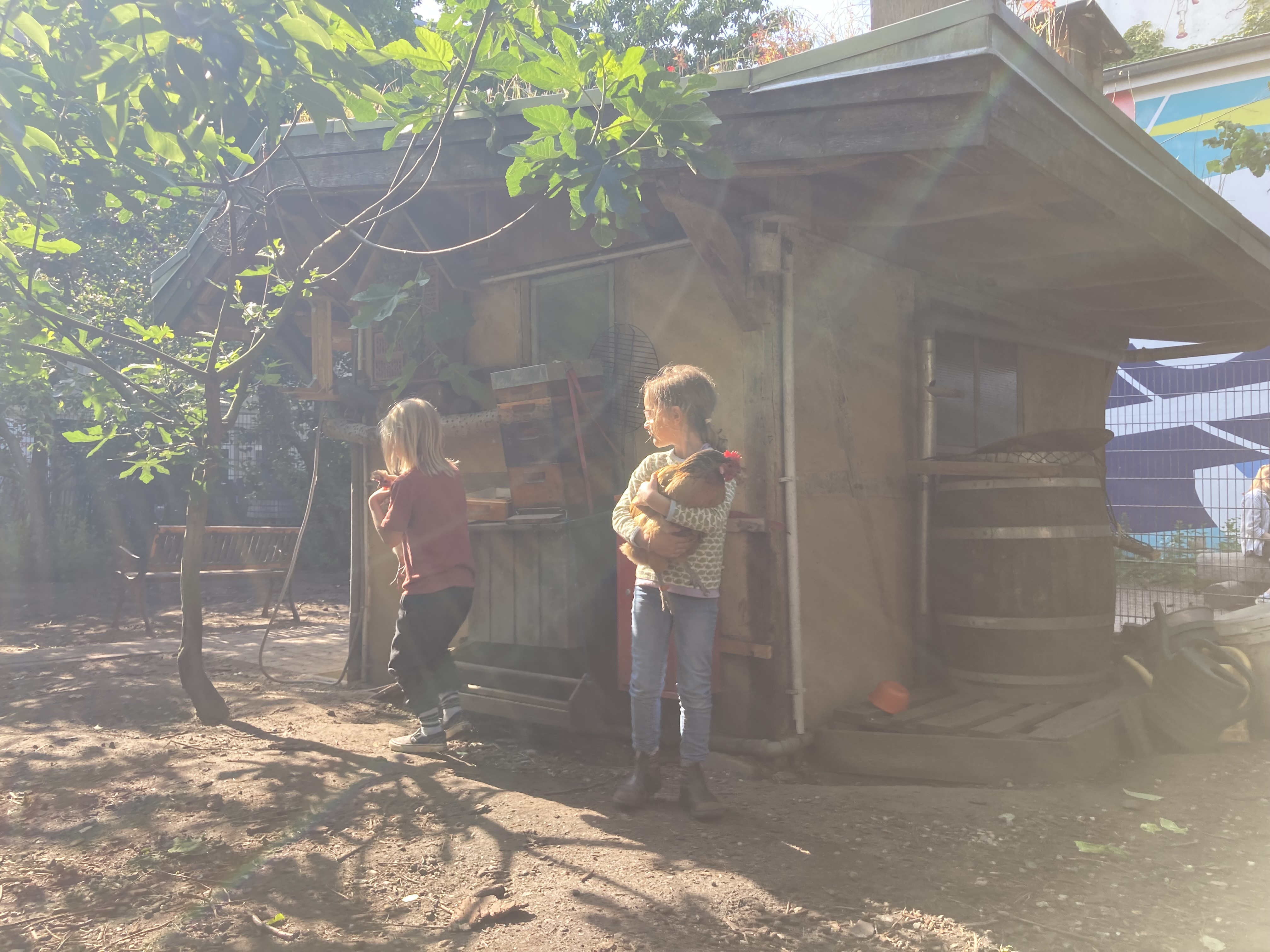 Zwei Kinder stehen im Garten neben einer Holzhütte, eines hält einen kleinen Hund, während Sonnenlicht durch Blätter fällt.