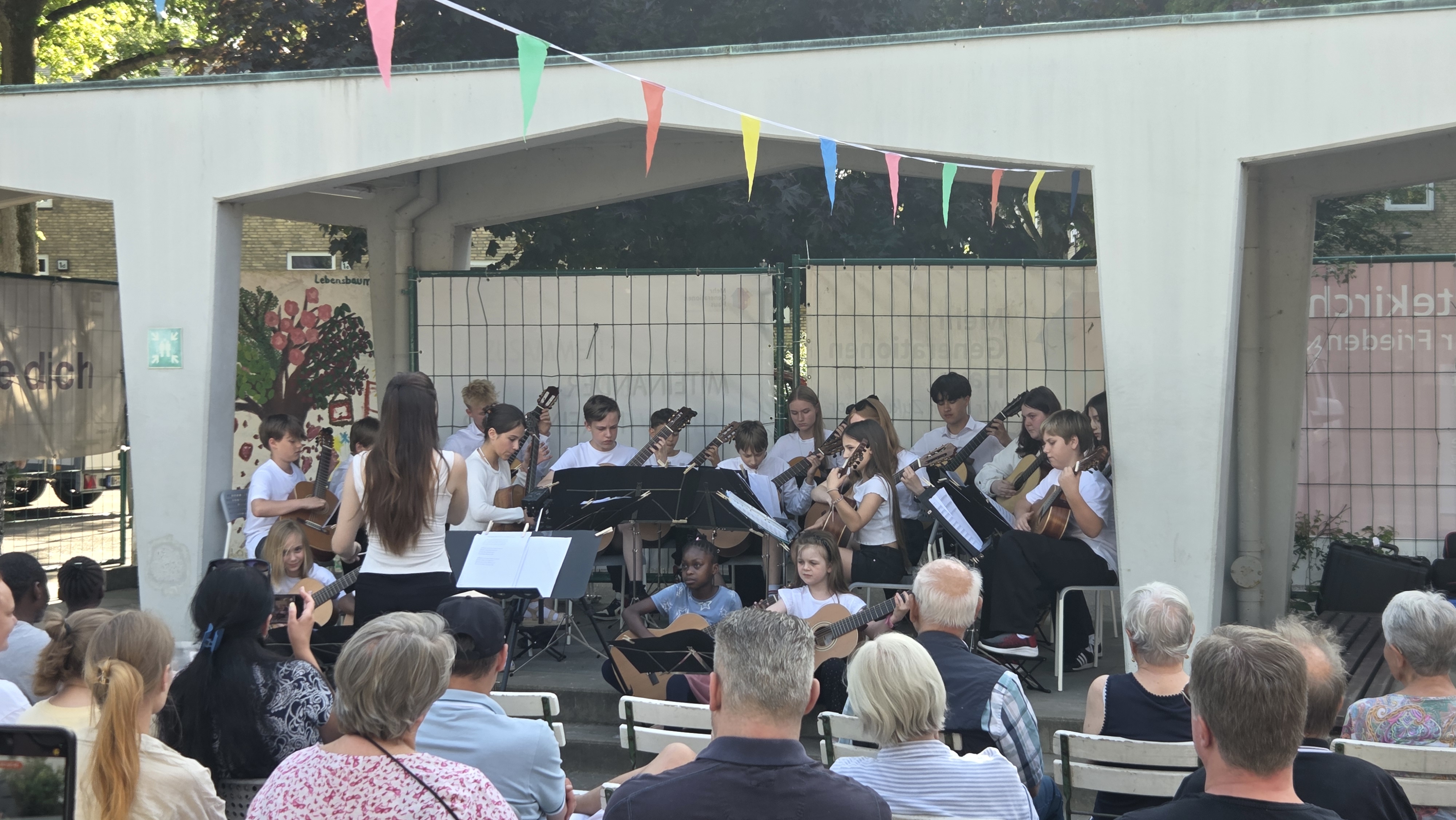 Jugendorchester mit Gitarren spielt draußen auf einer Bühne; bunte Fähnchen hängen darüber, Publikum schaut zu.
