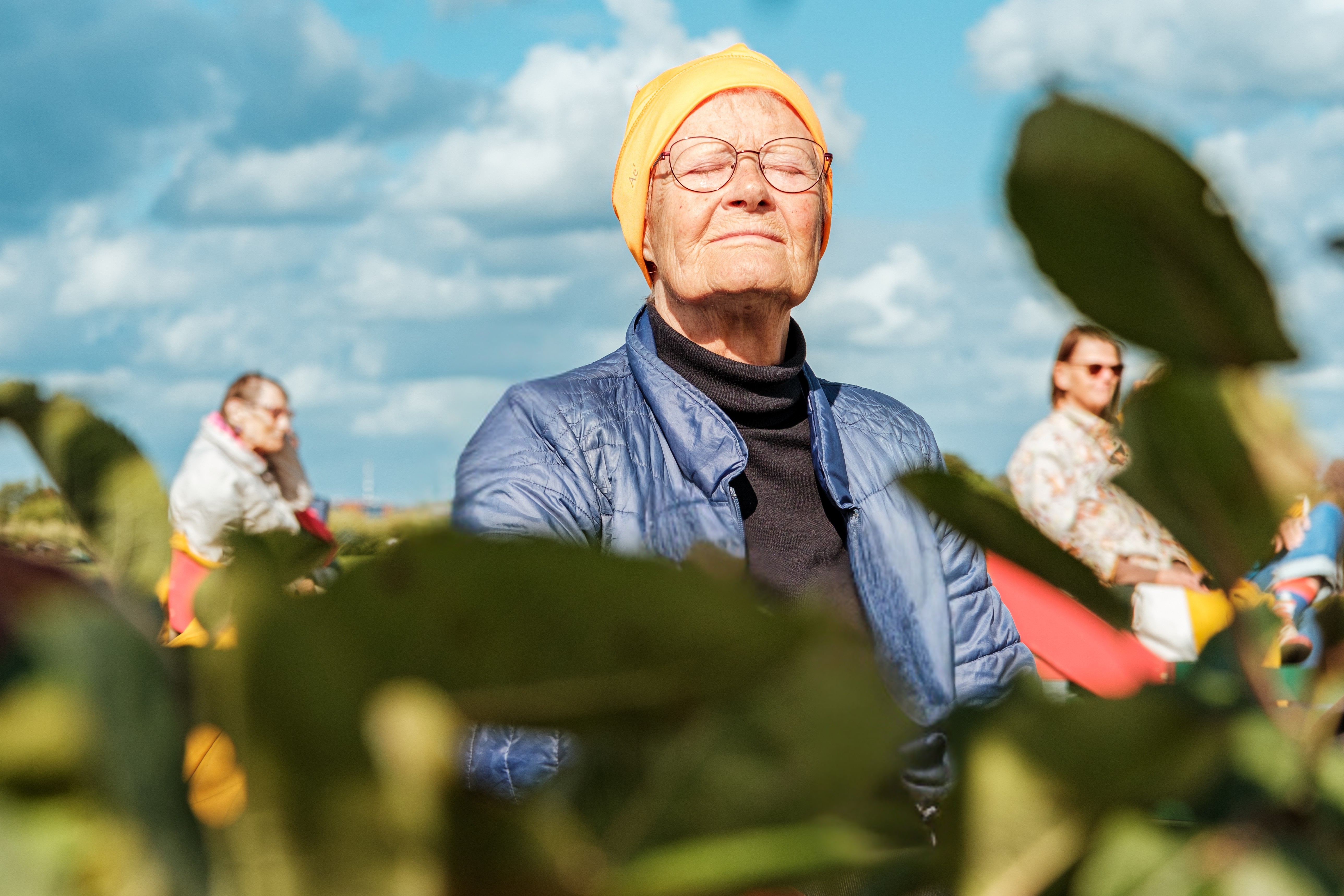 Ältere Frau mit orangefarbener Badekappe und Brille steht draußen, blickt nach oben, Himmel mit Wolken im Hintergrund.