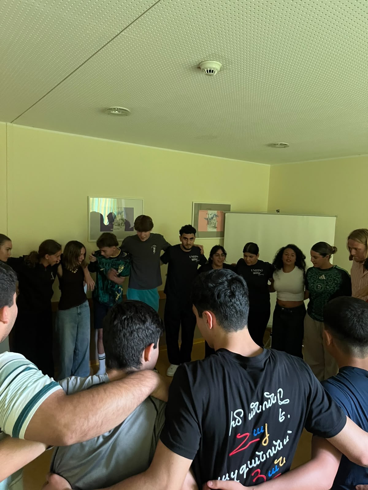 Gruppe junger Menschen steht im Kreis mit Armen um die Schultern in einem hellen Raum.