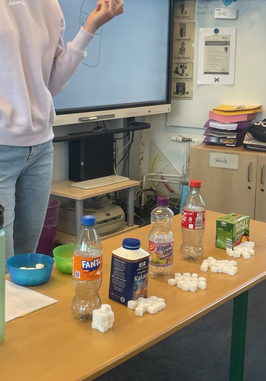 Eine Person steht links vor dem Smartboard; auf dem Tisch stehen Getränkeflaschen und Zuckerwürfel in Gruppen.