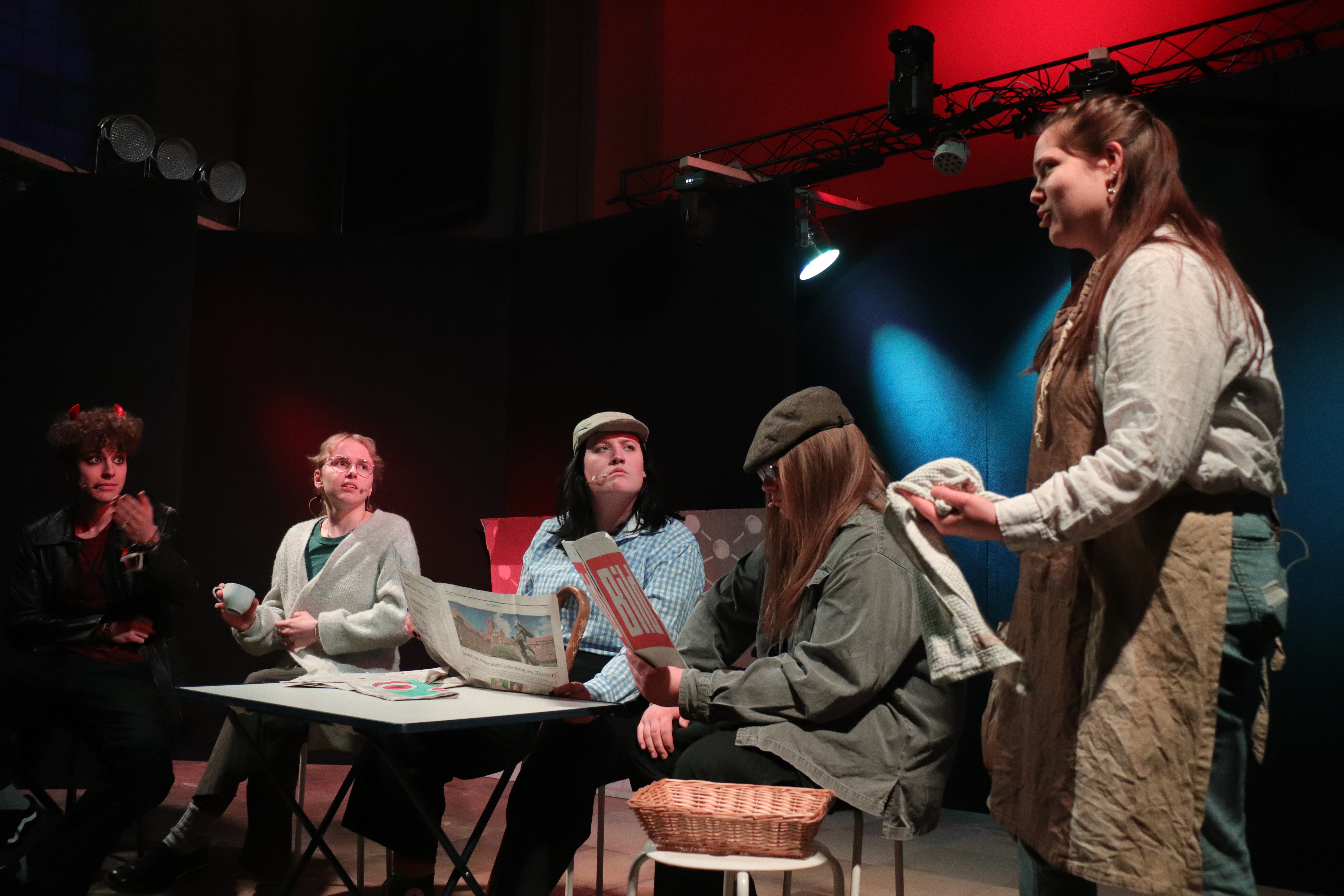 Gruppe von Schauspielern in Kostümen sitzt um einen Tisch mit Zeitungen auf einer Bühne; eine Frau steht rechts und spricht.