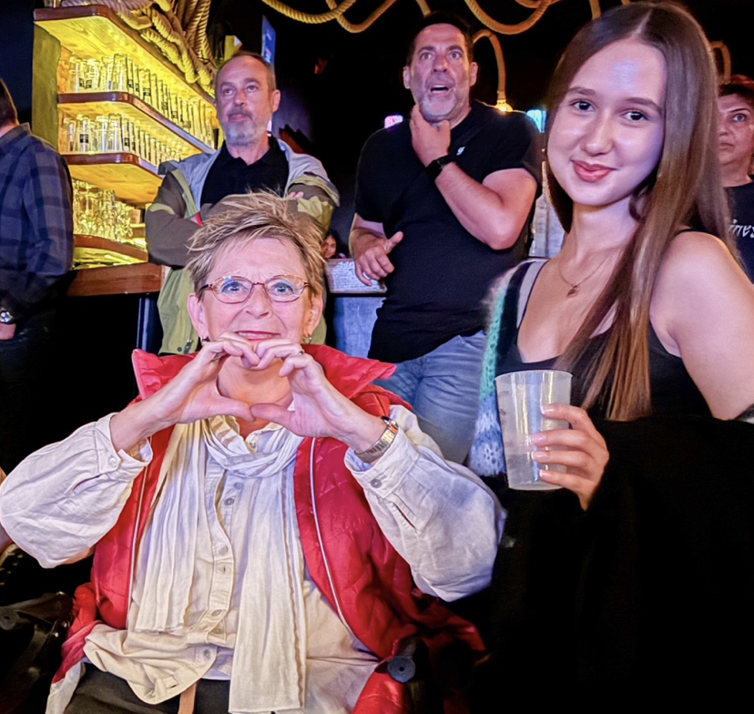 Ältere Frau mit Brille sitzt vorne, formt Herz mit den Händen; daneben junge Frau mit Getränk, Bar-Hintergrund.