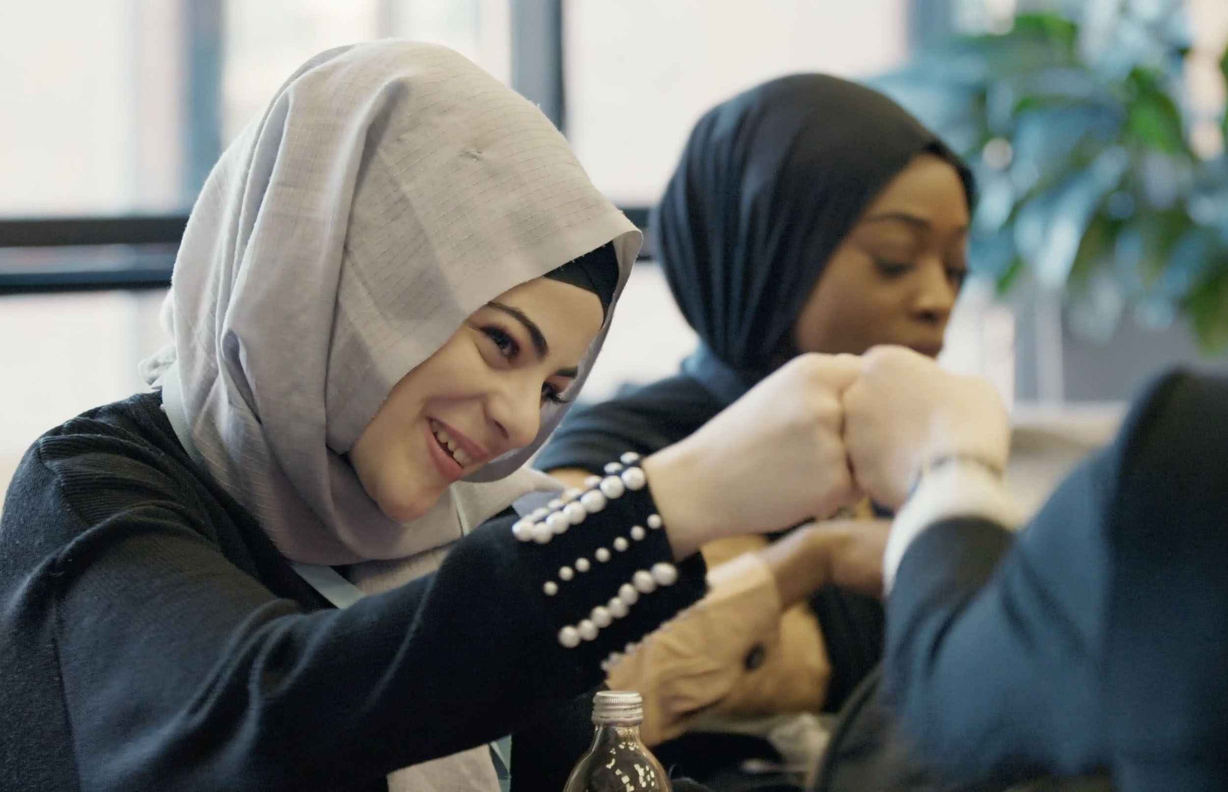 Zwei Frauen mit Kopftüchern sitzen am Tisch; die vordere lächelt in die Kamera, im Hintergrund eine Pflanze zu sehen.