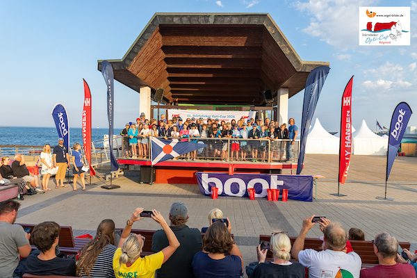 Gruppe von Menschen steht auf einer Bühne am Strand unter einem Holzdach; Publikum davor, Banner und Flaggen, Meer im Hinterg